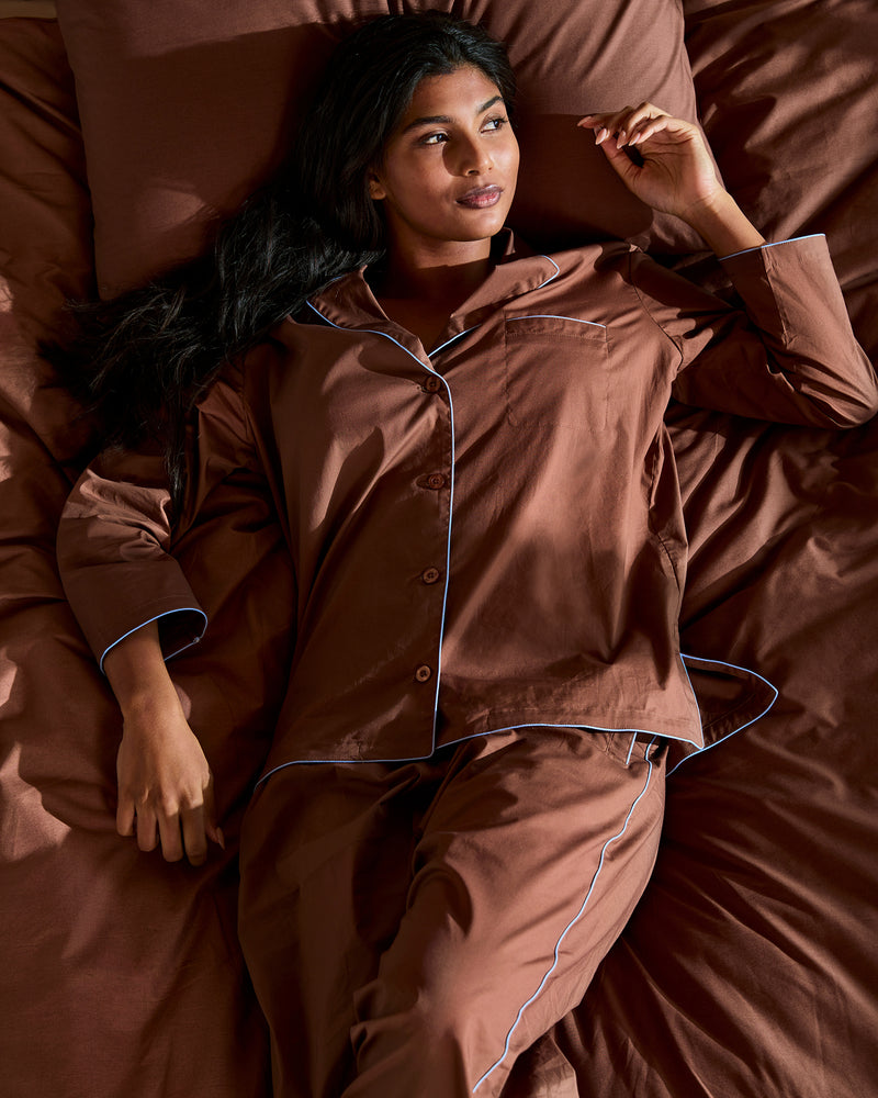 A woman in the Bed Threads Espresso & Sky Piped Long Sleep Set stands beside a bed with brown bedding in a sunlit room, next to a window with beige curtains and a brown lamp on the nightstand.