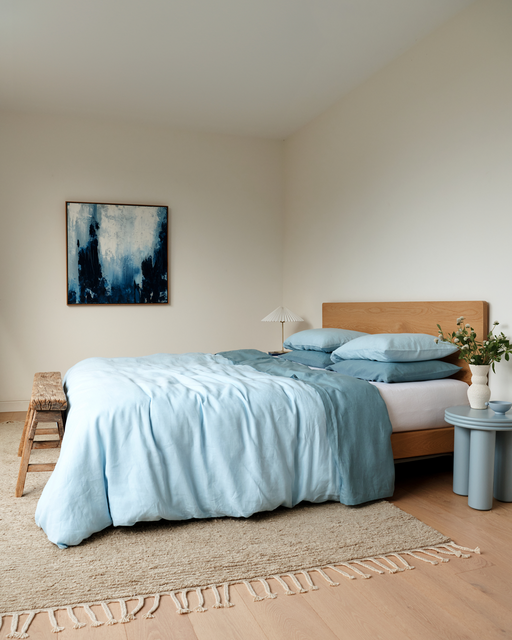 A cozy bedroom with a wooden bed frame, Bed Threads' Coast, Lagoon & White 100% French Flax Linen Bedding Bundle, a beige rug, small wooden bench, two blue side tables with a plant and lamp, plus an abstract blue and white wall painting.
