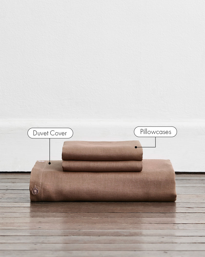A folded duvet cover in a warm taupe-brown colour called Portobello in 100% french flax linen, sits on a wooden floor against a white wall. Two stacked pillowcases in the same shade sit on top of the duvet. Two callout labels point to the stacked duvet cover and pillowcases, identifying the product components.