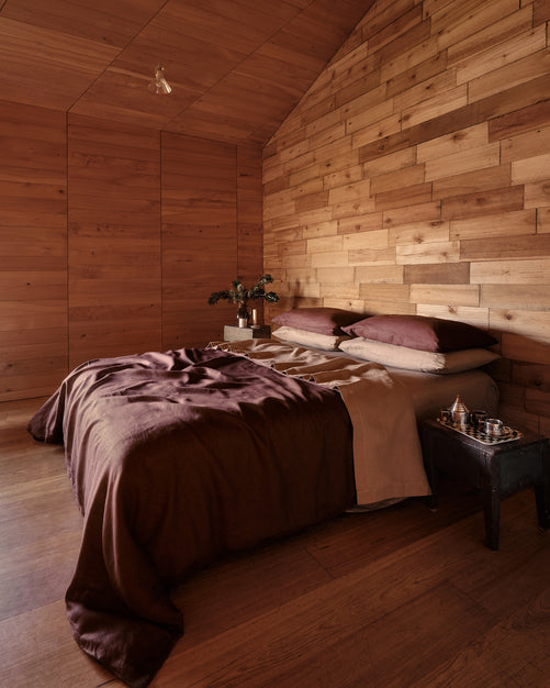 A cozy bedroom with a bed dressed in cacao and portobello 100% french flax linen bedding, set against a wall and ceiling made of warm wood paneling. A small bedside table holds a glass and a small decorative object.