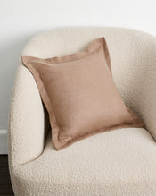 A square warm taupe cushion cover in 100% french flax linen with a subtle border detail, resting on a textured cream boucle armchair against a plain white wall. - view