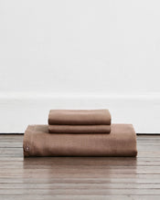 A folded duvet cover in a warm taupe-brown colour called Portobello in 100% french flax linen, sits on a wooden floor against a white wall. Two stacked pillowcases in the same shade sit on top of the duvet. - view