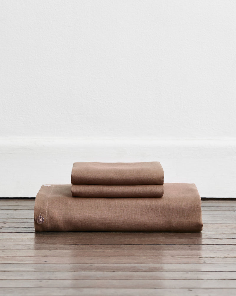A folded duvet cover in a warm taupe-brown colour called Portobello in 100% french flax linen, sits on a wooden floor against a white wall. Two stacked pillowcases in the same shade sit on top of the duvet.