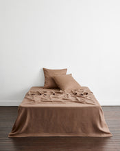 A bed sits in a room with dark wooden floors and a white wall, dressed in warm taupe-brown 100% french flax linens. A flat sheet drapes onto the floor and two european pillowcases lean against the white wall. - view