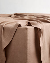 A close-up of a draped taupe-brown flat sheet in 100% french flax linen resting on the corner of a fitted sheet, showing its soft texture and folds against a neutral light background. - view