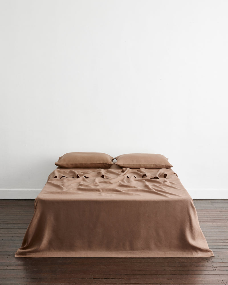 A bed dressed in a smooth, taupe-brown 100% french flax linen sheet and two matching pillows, styled with a ruffled sheet detail on the top, set against a minimalist white wall and dark wood floor.
