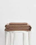 A stack of folded brown 100% french flax linens, including a fitted sheet, flat sheet and pillowcase cover, displayed on a white stool against a plain white background.