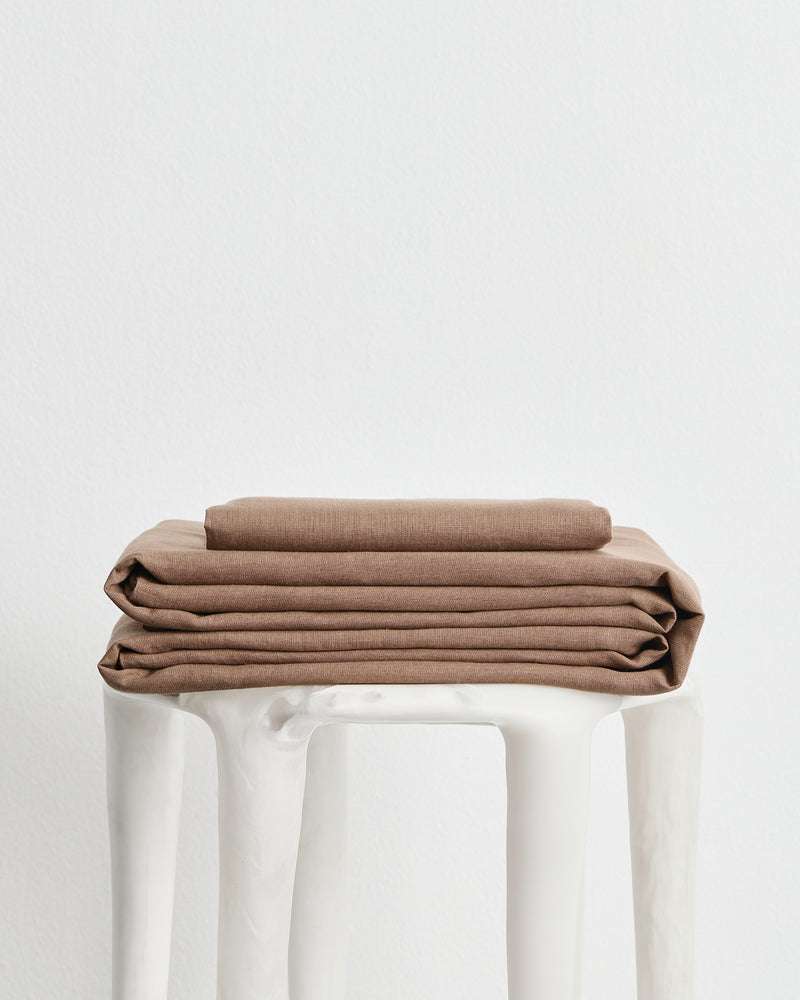 A stack of folded brown 100% french flax linens, including a fitted sheet, flat sheet and pillowcase cover, displayed on a white stool against a plain white background.