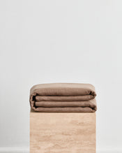A folded warm brown throw in 100% french flax linen, folded neatly on a light beige stone pedestal against a plain white wall. - view