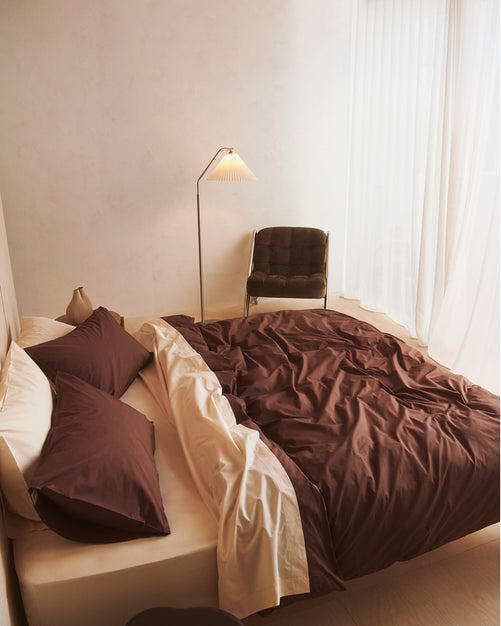 A bed styled with the Bed Threads Espresso & Vanilla Organic Cotton Bedding Bundle shows cream bedding and dark brown accents. Pillowcases and a matching percale duvet cover rest atop, while a bedside table holds a vase next to a round table in view.