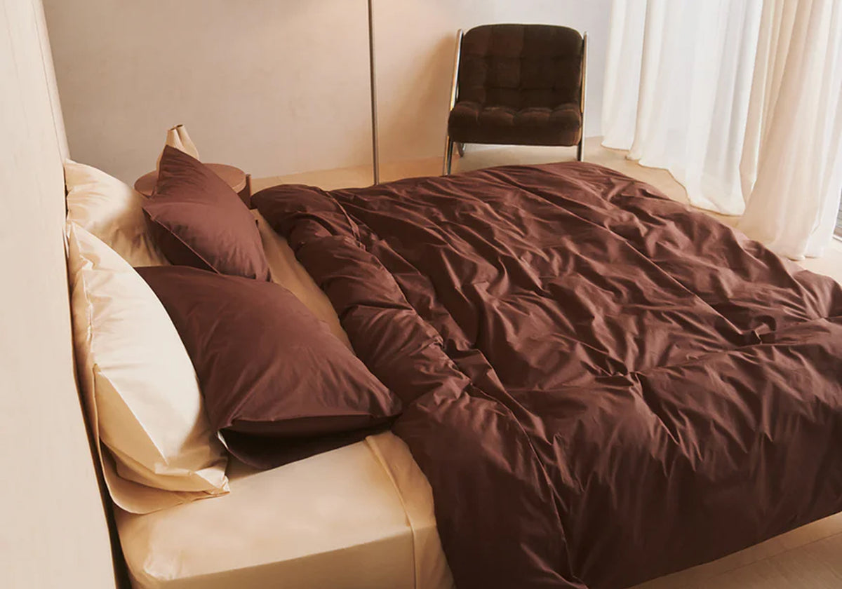 A neatly made bed with cream-colored sheets and brown bedding is positioned near a window with sheer white curtains. A dark upholstered chair sits in the background beside the bed.