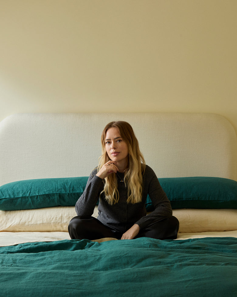 A woman with long blonde hair sits on a neatly made bed with teal bedding, leaning slightly forward and resting her hand near her chin, against a light-colored, upholstered headboard in a softly lit room.