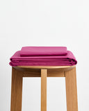 A bed with two pillows and wrinkled Bed Threads Berry Organic Cotton Fitted Sheet, GOTS certified percale in solid bright pink, set against a plain white wall.