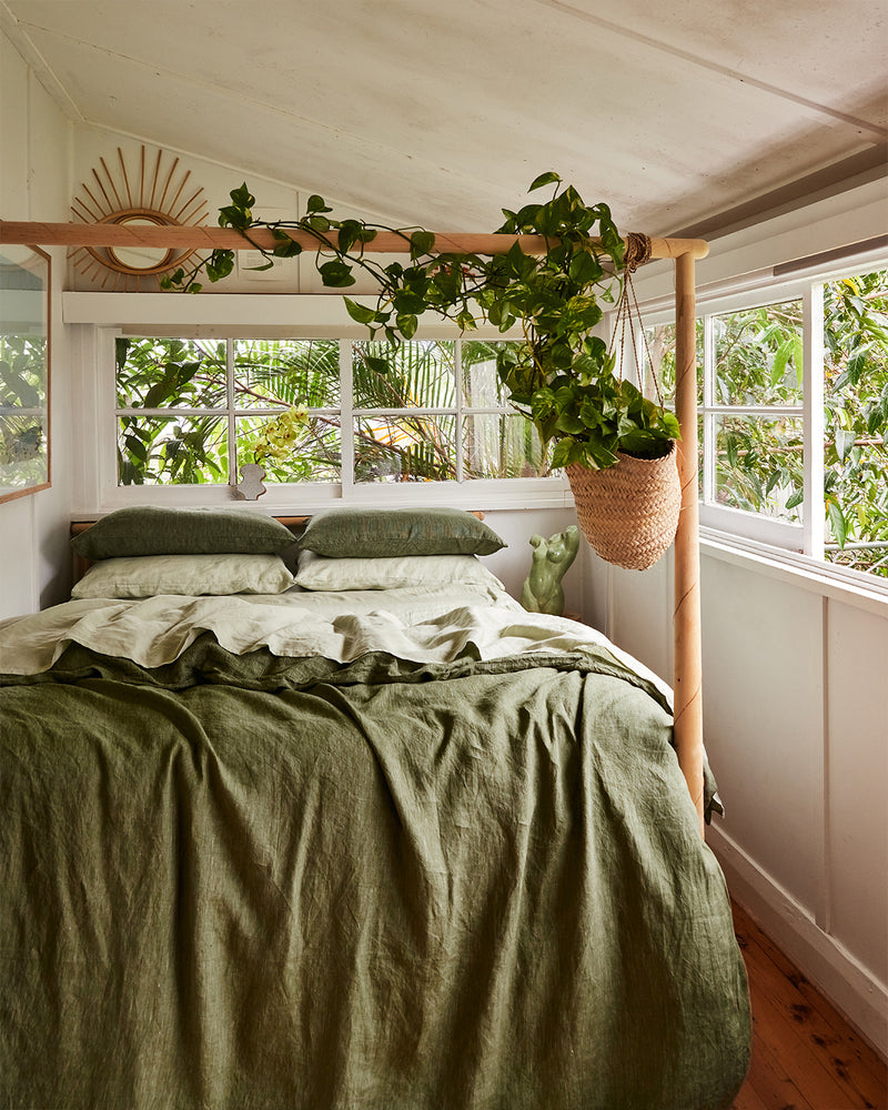 A cozy bedroom features a canopy bed dressed in the Bed Threads Sage 100% French Flax Linen Fitted Sheet, soft pillows, and a hanging potted plant. Large windows fill the white-walled, wood-floored room with light and views of lush greenery.