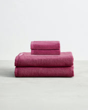 Set of two berry pink face washers and two hand towels in 100% cotton terry stacked on a light grey surface with a white background.  - view