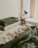 A neatly made bed features the Bed Threads Olive & Oatmeal & White Stripe 100% French Flax Linen Bedding Bundle, complete with matching pillows and duvet. A bedside table holds a lamp and vase with purple flowers, while soft light filters through neutral curtains.