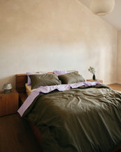 A bed dressed in olive green bedding with violet and vanilla sheets, styled in a minimalist bedroom with wooden nightstands and a textured plaster wall. - view