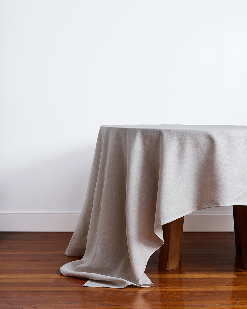 An Oatmeal 100% French Flax Linen Tablecloth by Bed Threads draped over a wooden table, set against a plain white wall and hardwood floor.