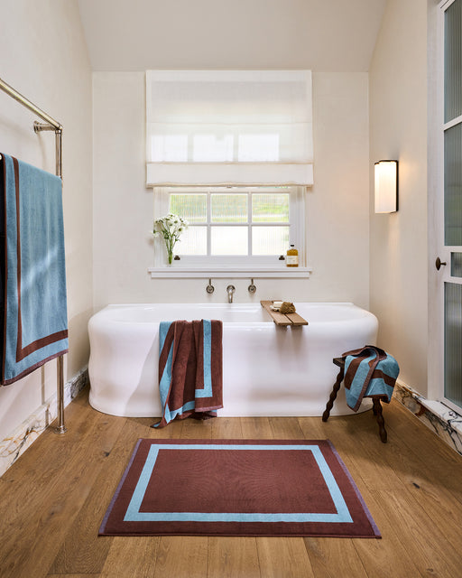 A woman sits on the edge of a white bathtub, wrapped in a Bed Threads Cacao & Lagoon Two-Toned 100% Cotton Terry Bath Towel, with a matching towel on her head. A rug, stool with toiletries, and fresh flowers complete the scene.