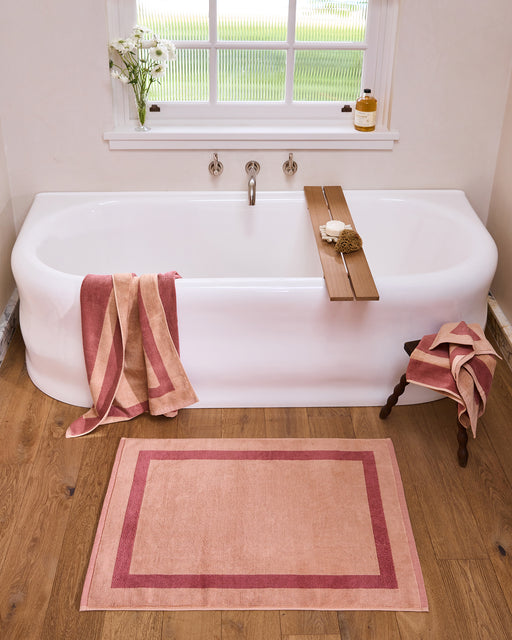 A woman wrapped in a Bed Threads Terracotta & Pink Clay Two-Toned 100% Cotton Terry Bath Towel stands in a modern bathroom with marble counters, a vase of flowers, and matching towels and mat beside a glass shower.