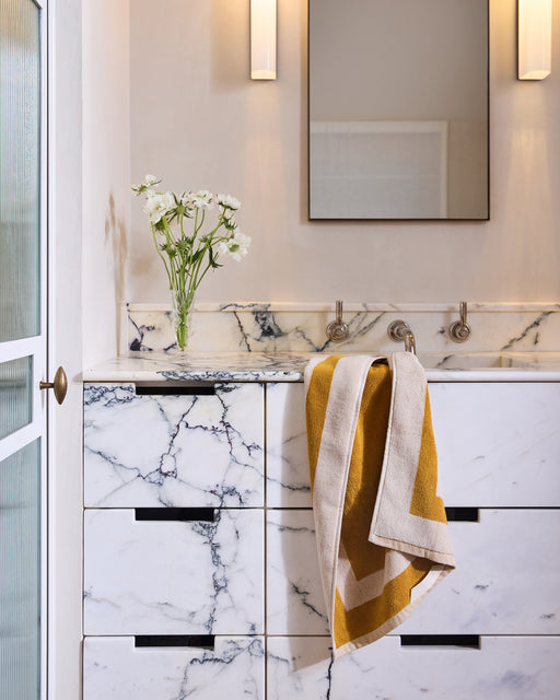 Wearing a beige bathrobe, a person stands by the tub, holding two Bed Threads Crème & Turmeric Two-Toned 100% Cotton Terry Hand Towels as sunlight pours through the window.