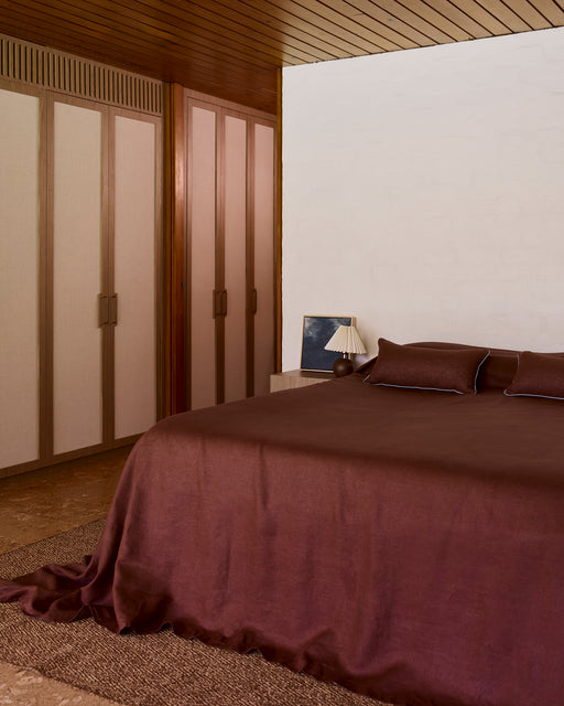 A neatly made bed features dark brown heavy linen bedding and the Bed Threads Cacao & Lagoon 100% French Flax Linen Piped Lumbar Cushion Cover. Beside it, a wooden nightstand holds a pleated lamp and a framed picture with a dark image.