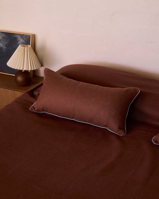 A neatly made bed features dark brown heavy linen bedding and the Bed Threads Cacao & Lagoon 100% French Flax Linen Piped Lumbar Cushion Cover. Beside it, a wooden nightstand holds a pleated lamp and a framed picture with a dark image.