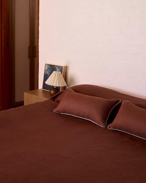 A neatly made bed with Bed Threads' Cacao & Lagoon 100% French Flax Linen Piped Lumbar Cushion Cover and matching pillows is beside a wooden nightstand with a pleated lampshade and framed photo, set against a white brick wall.