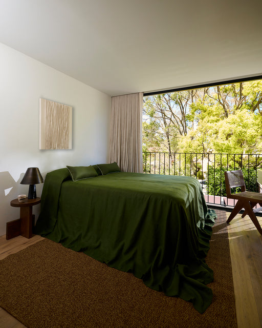 A modern bedroom featuring the Bed Threads Olive Heavy Linen Bed Cover, beige curtains, a large window, and a view of leafy green trees, filling the space with natural sunlight.