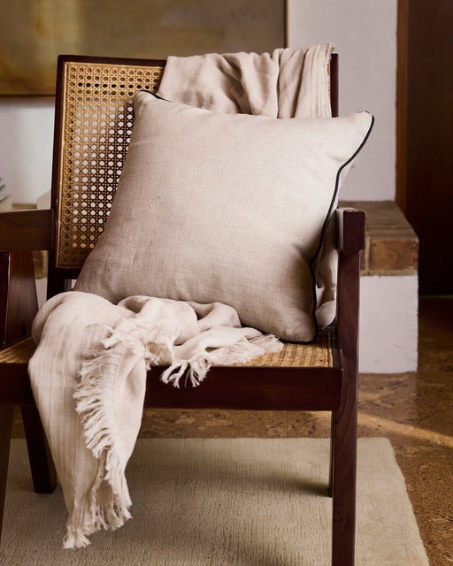 A wooden chair with a woven cane seat is draped with a light beige throw and the Bed Threads Oatmeal & Charcoal 100% French Flax Linen Piped Cushion Cover, set in a cozy, softly lit room with textured floors and neutral decor.