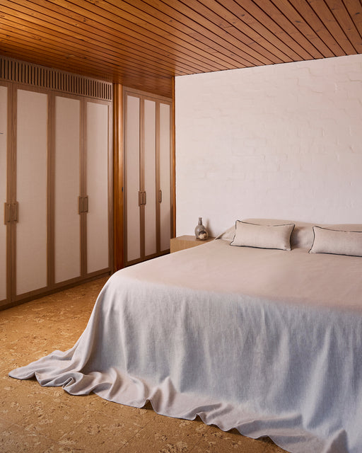 A neatly made bed with beige heavy linen and two Bed Threads Oatmeal & Charcoal 100% French Flax Linen Piped Lumbar Cushion Covers sits by a wooden nightstand with a small vase and decor, against a light-colored wall.