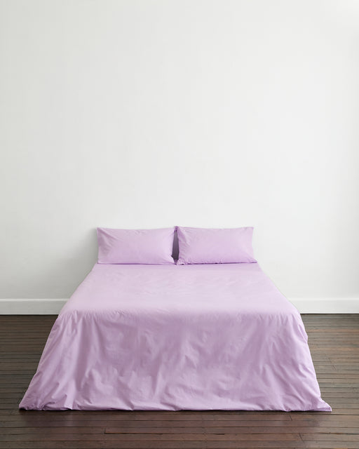 A neatly made bed with the Bed Threads Violet 100% Organic Cotton Duvet Cover and cream pillows sits in a bright, minimalist bedroom with light wood floors, a large paper lantern ceiling light, and soft natural light through sheer curtains.