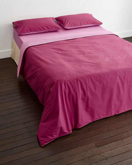 A bed is styled with Bed Threads’ Berry, Bubblegum & Moss Piped 100% Organic Cotton Duvet Cover, two matching pillows, and a light pink sheet, all arranged on a dark wooden floor beside a plain white wall.