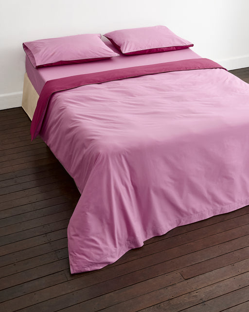 A neatly made bed with light pink sheets, two pillows, and the Bed Threads Berry, Bubblegum & Moss Piped 100% Organic Cotton Duvet Cover in a percale weave, set on a dark wooden floor against a white wall.