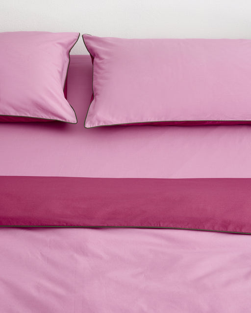 A neatly made bed featuring Bed Threads’ Berry, Bubblegum & Moss Piped 100% Organic Cotton Duvet Cover with two pink pillows and a darker pink strip at the foot, set against a plain white wall.