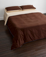 A neatly made bed with dark brown sheets, beige top sheet, and Bed Threads' Espresso, Vanilla & Sky Piped 100% Organic Cotton Duvet Cover sits against a white wall on a dark wooden floor. - view
