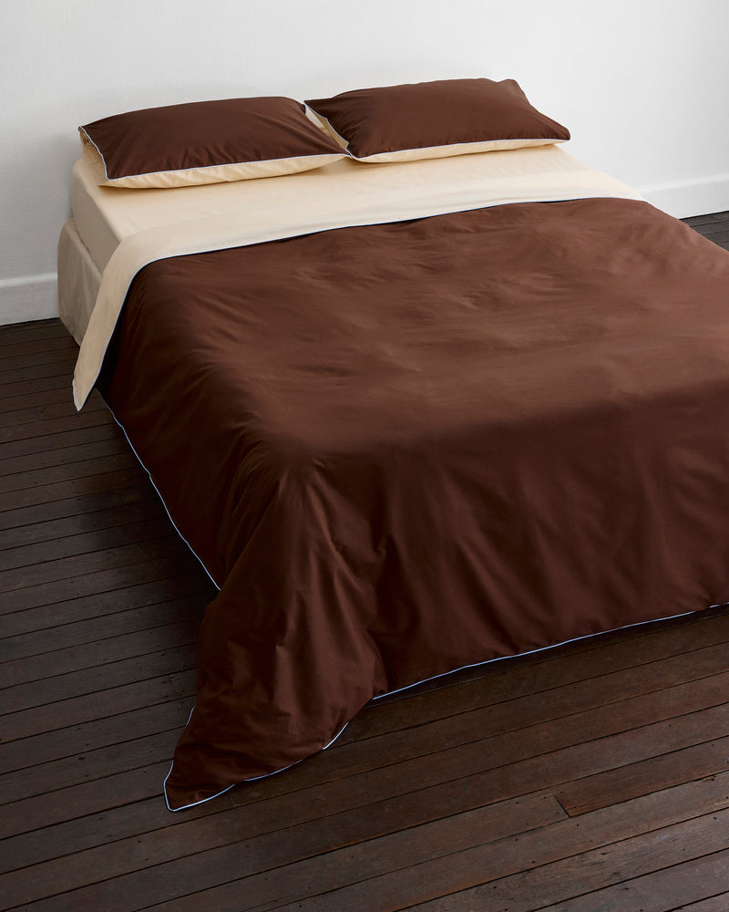 A neatly made bed with dark brown sheets, beige top sheet, and Bed Threads' Espresso, Vanilla & Sky Piped 100% Organic Cotton Duvet Cover sits against a white wall on a dark wooden floor.