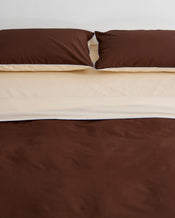 A neatly made bed features two brown pillows and the Bed Threads Espresso, Vanilla & Sky Piped 100% Organic Cotton Duvet Cover, layered over a cream sheet, set against a plain white wall. - view