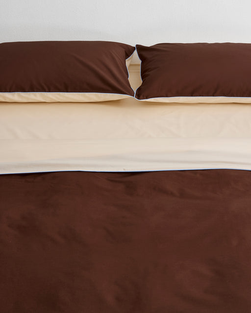 A neatly made bed features two brown pillows and the Bed Threads Espresso, Vanilla & Sky Piped 100% Organic Cotton Duvet Cover, layered over a cream sheet, set against a plain white wall.