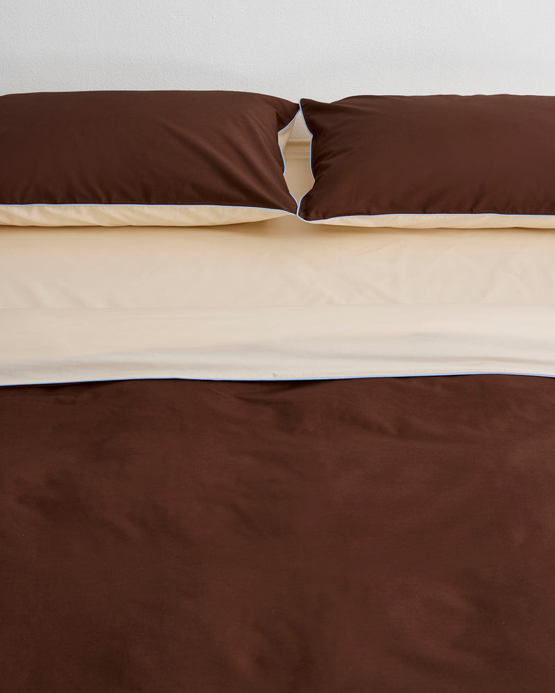 A neatly made bed features two brown pillows and the Bed Threads Espresso, Vanilla & Sky Piped 100% Organic Cotton Duvet Cover, layered over a cream sheet, set against a plain white wall.