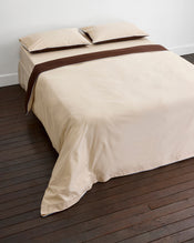 A neatly made bed with cream-colored sheets and pillowcases, topped with a brown blanket, features Bed Threads' Espresso, Vanilla & Sky Piped 100% Organic Cotton Duvet Cover on a dark wooden floor against a plain white wall. - view