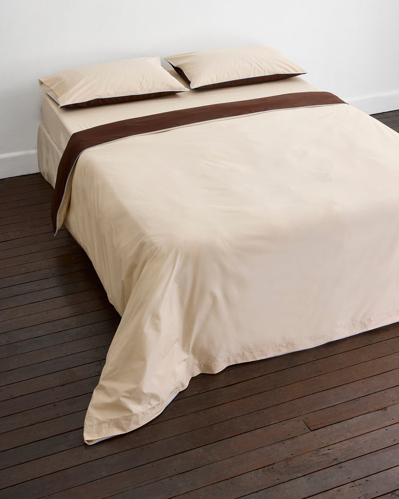 A neatly made bed with cream-colored sheets and pillowcases, topped with a brown blanket, features Bed Threads' Espresso, Vanilla & Sky Piped 100% Organic Cotton Duvet Cover on a dark wooden floor against a plain white wall.
