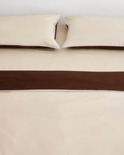 A neatly made bed with beige sheets and two matching pillows, featuring a wide dark brown bed runner, is complemented by the Bed Threads Espresso, Vanilla & Sky Piped 100% Organic Cotton Duvet Cover. - view