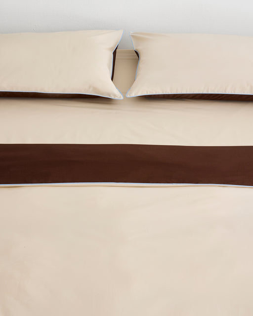 A neatly made bed with beige sheets and two matching pillows, featuring a wide dark brown bed runner, is complemented by the Bed Threads Espresso, Vanilla & Sky Piped 100% Organic Cotton Duvet Cover.
