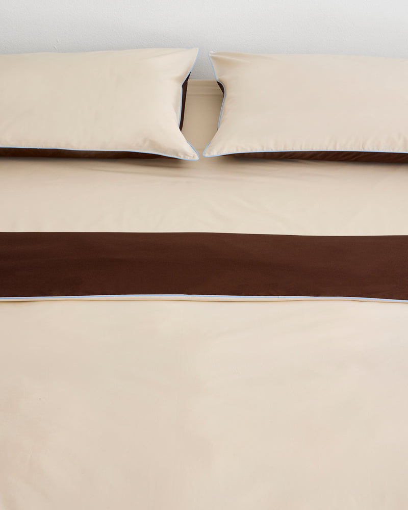 A neatly made bed with beige sheets and two matching pillows, featuring a wide dark brown bed runner, is complemented by the Bed Threads Espresso, Vanilla & Sky Piped 100% Organic Cotton Duvet Cover.