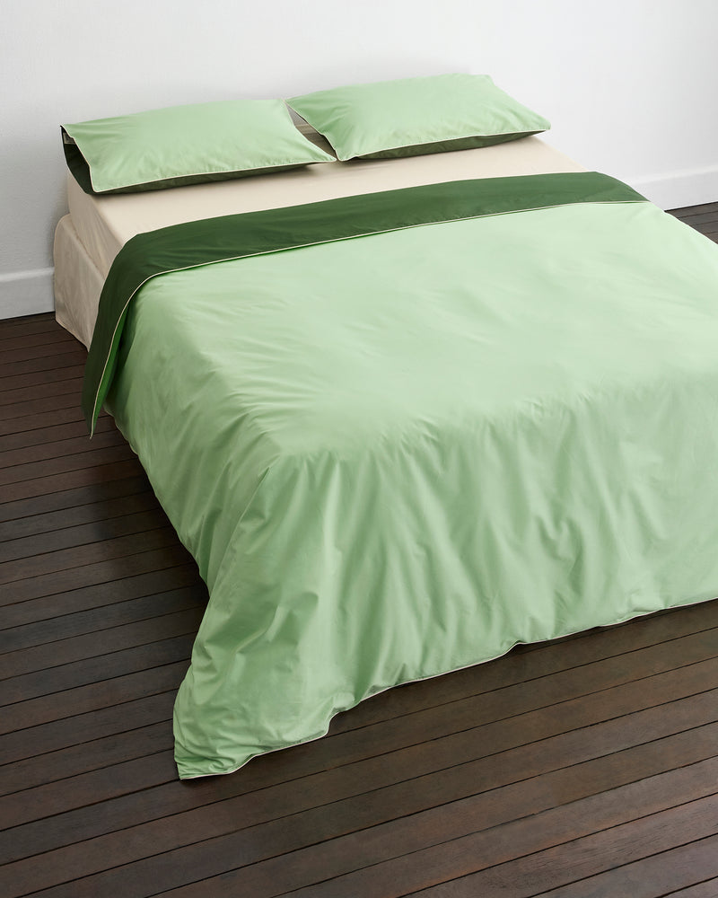 A neatly made bed with light green sheets, matching pillowcases, and a dark green accent strip across the top, set on dark wooden flooring against a white wall.