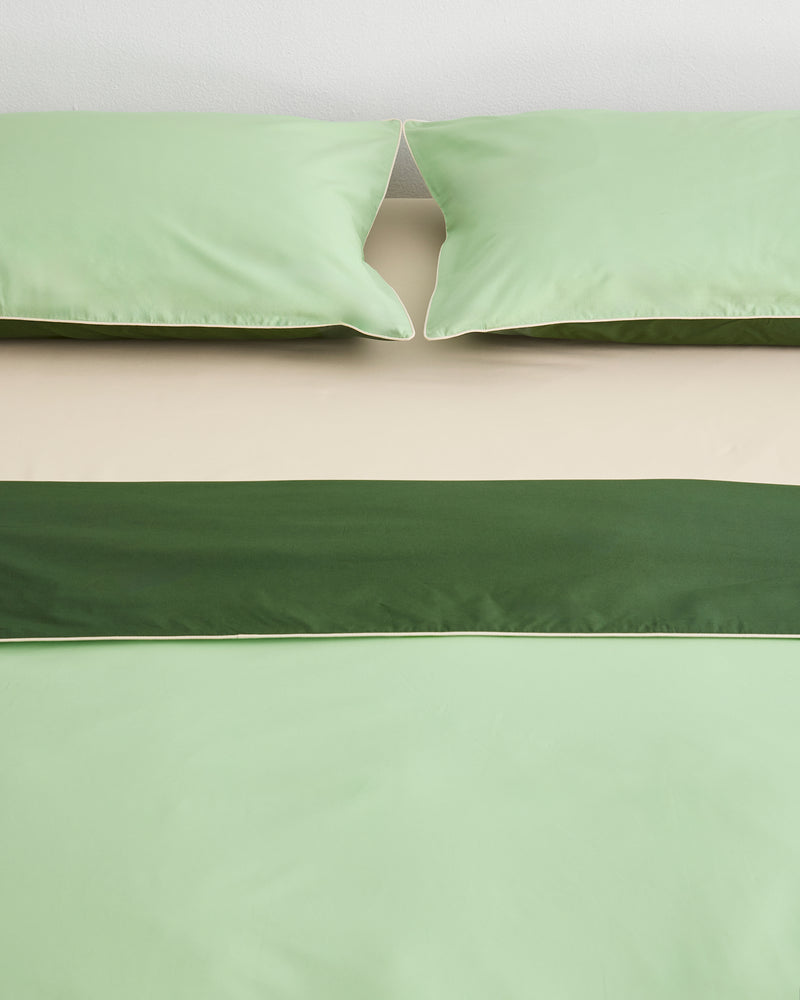 A neatly made bed with light green pillows and bedding, a beige sheet, and a dark green decorative band across the bed. The scene is minimal and the colors are soft and calming.