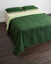 A neatly made bed with two green pillows, a light green top sheet, and a dark green Moss, Apple & Vanilla Piped 100% Organic Cotton Duvet Cover by Bed Threads, set against a white wall on a dark wooden floor. - view