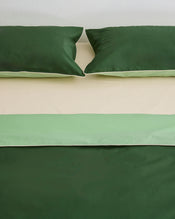Two green pillows sit on a beige bed dressed in the Bed Threads Moss, Apple & Vanilla Piped 100% Organic Cotton Duvet Cover. The percale bedding is neatly arranged against a plain white wall for a fresh, inviting look. - view