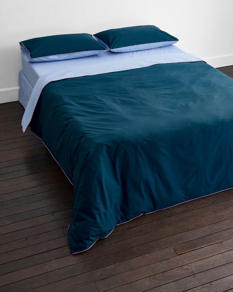 A neatly made bed features the Bed Threads Aegean, Sky & Bubblegum Piped 100% Organic Cotton Duvet Cover with matching dark blue percale bedding and pillows, all set against a white wall on a dark wooden floor.
